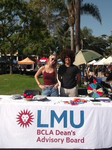 Two students standing behind a white tablecloth covered table outside on alumni mall at LMU